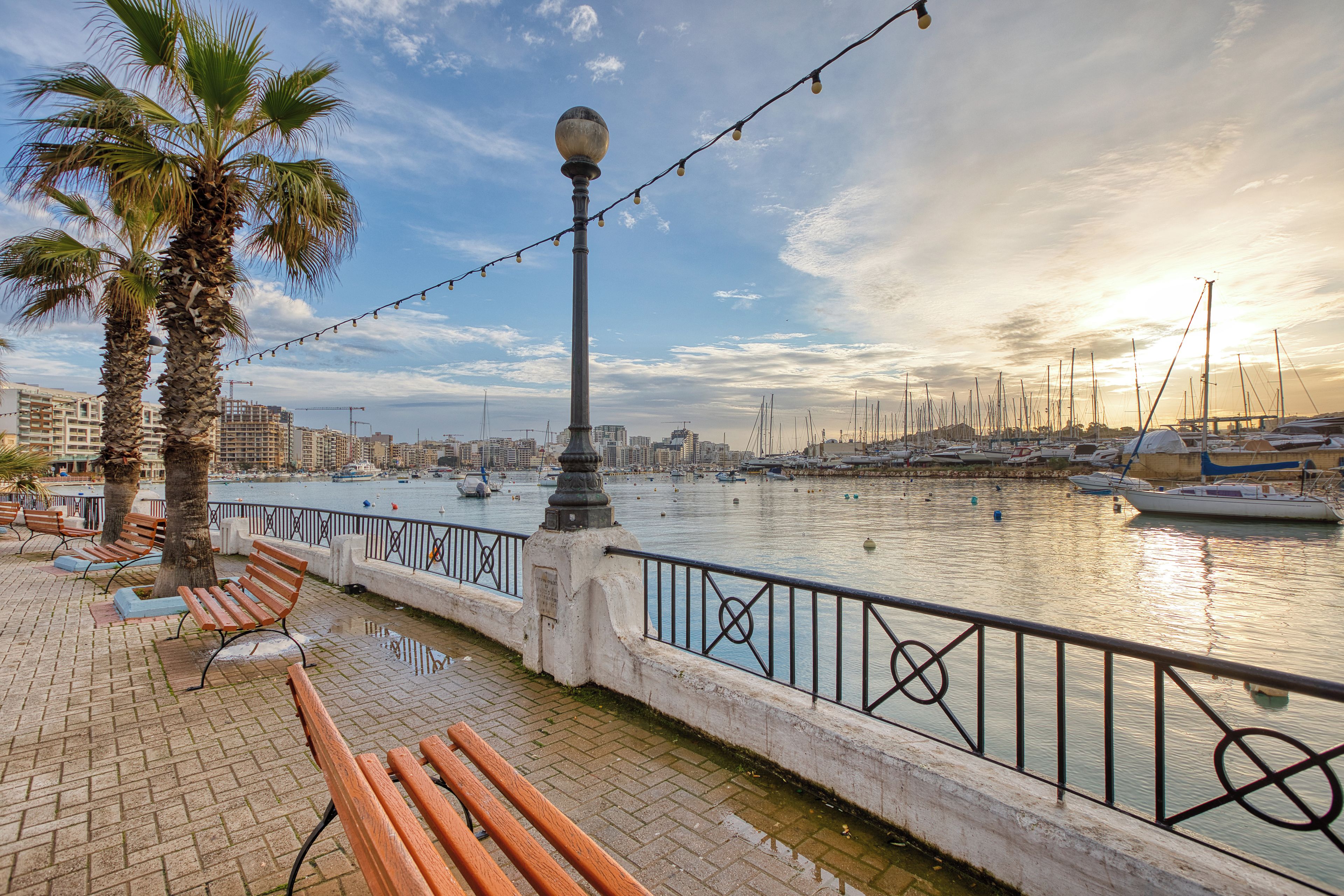 A waterfront promenade with benches, a lamp post, and a view of the marina with boats.