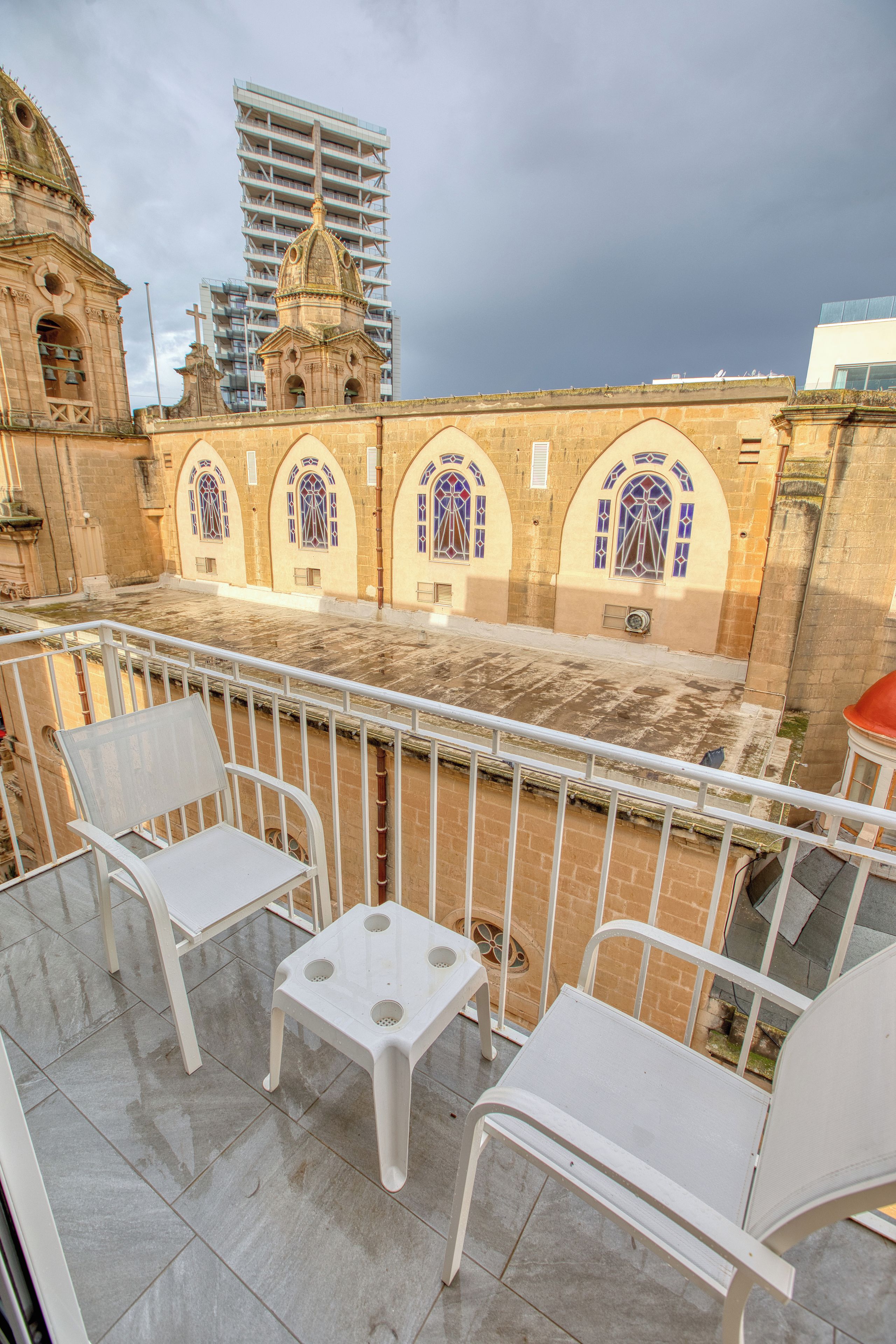 Un balcon avec des chaises blanches et une table, d’où l’on peut admirer un bâtiment historique doté d’arcades et d’un clocher.