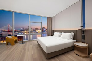 A modern hotel room with a large bed, a small round table, a yellow chair, and a view of a city skyline at dusk.
