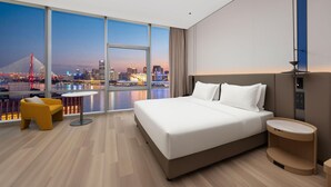 A modern hotel room with a large bed, a small round table, a yellow chair, and a view of a city skyline at dusk. - Holiday Inn Express Shanghai North Bund Riverside (Shanghai)