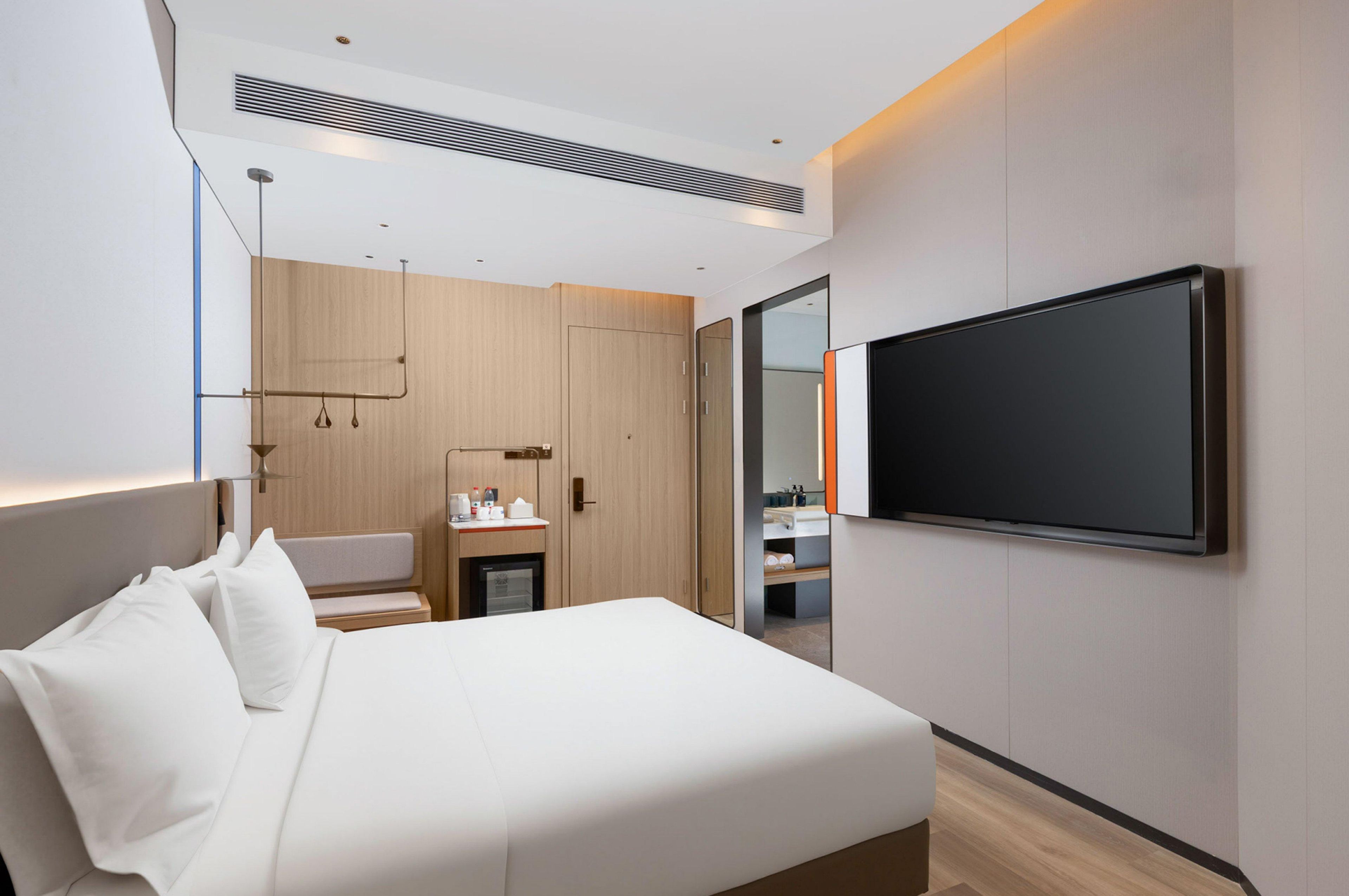A modern hotel room with a large bed, a flat-screen TV mounted on the wall, a small desk with a chair, and a wardrobe.