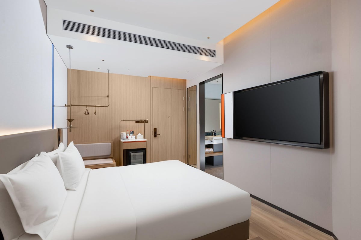 A modern hotel room with a large bed, a flat-screen TV mounted on the wall, a small desk with a chair, and a wardrobe.