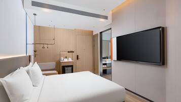 A modern hotel room with a large bed, a flat-screen TV mounted on the wall, a small desk with a chair, and a wardrobe.