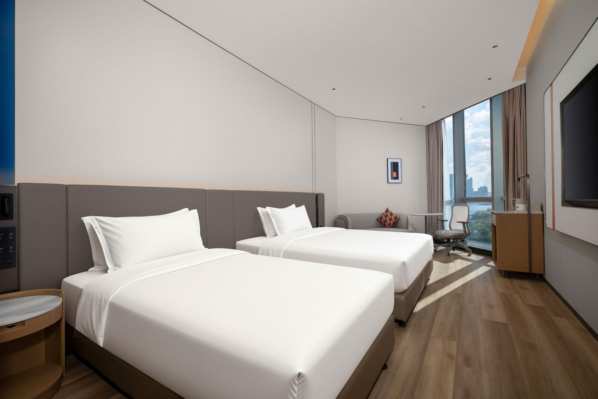 A hotel room with two beds, a desk, a chair, a TV, and a large window with a city view.