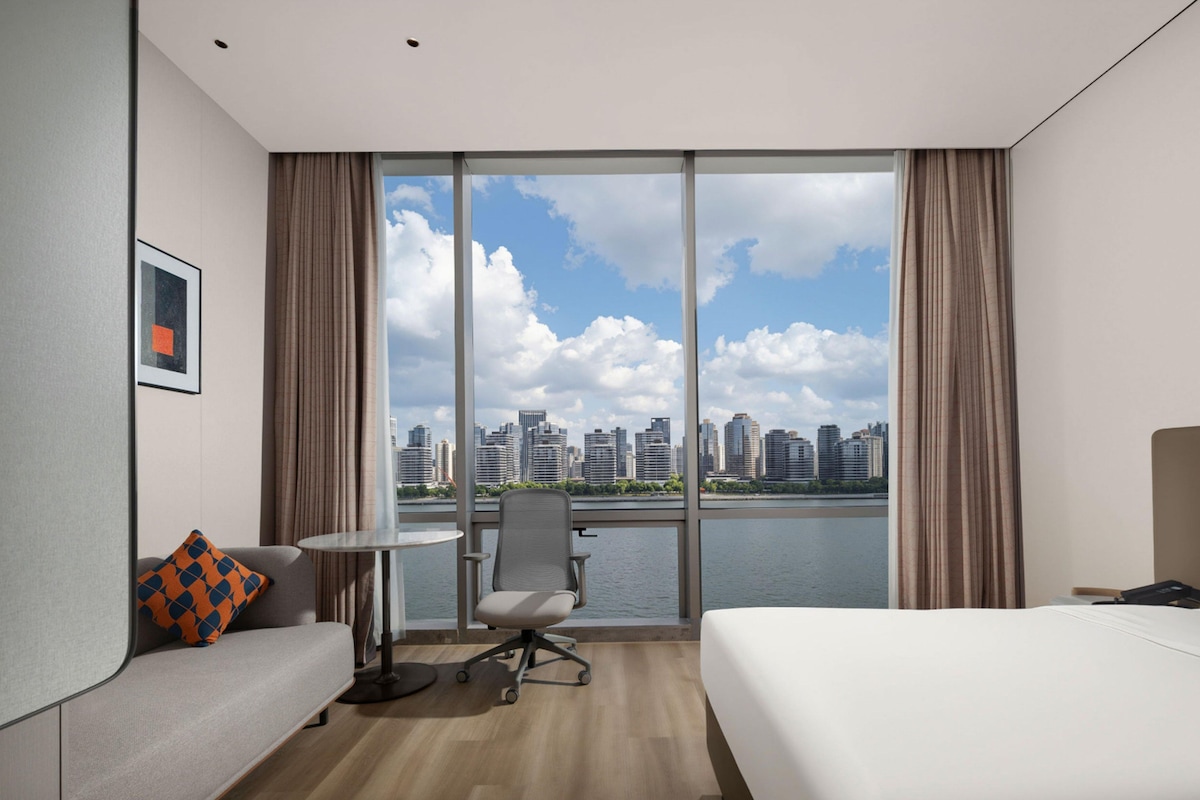 A hotel room with a bed, a desk, a chair, a sofa, and a large window with a city view.