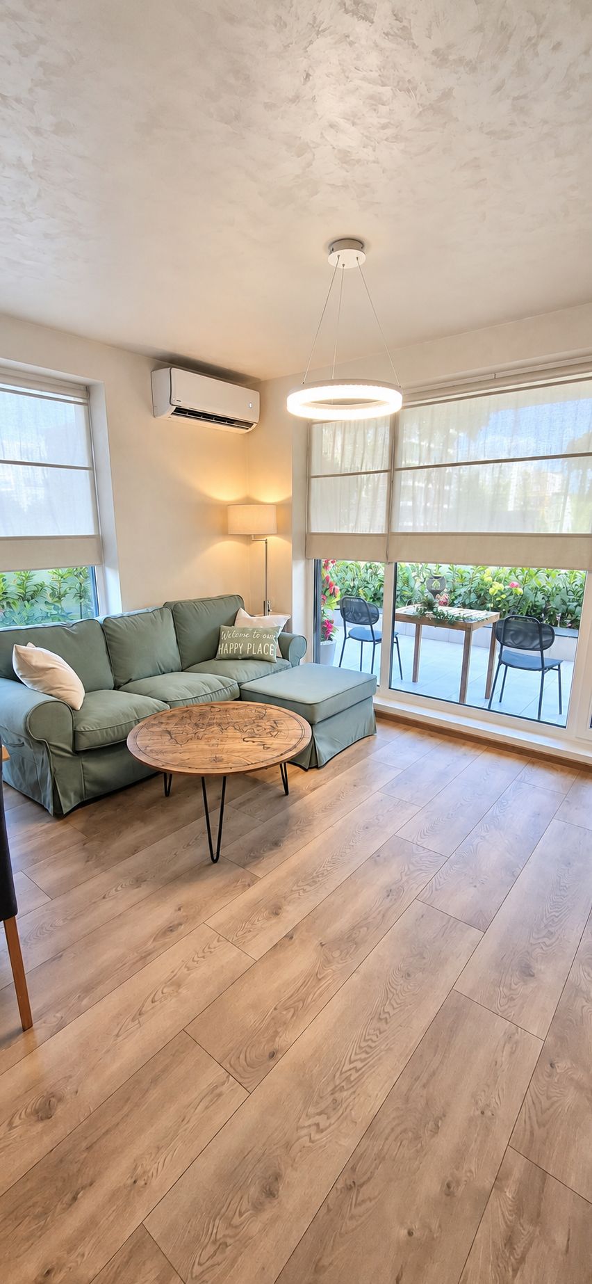 Een ruime woonkamer met een groene bank, een houten salontafel en uitzicht op groen door grote ramen.
