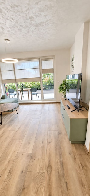Una habitación amplia con piso de madera, un sofá verde, un escritorio con televisor y ventanas grandes con vista a follaje.