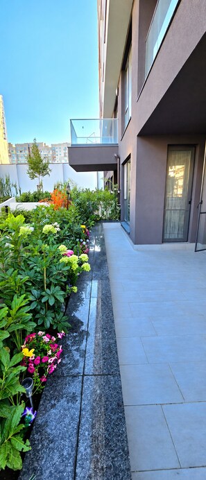 A modern building with a covered walkway, a garden with various plants and flowers, and a clear sky. - Stylish Apartment with Large Green Terrace & Private Parking (Sofia)