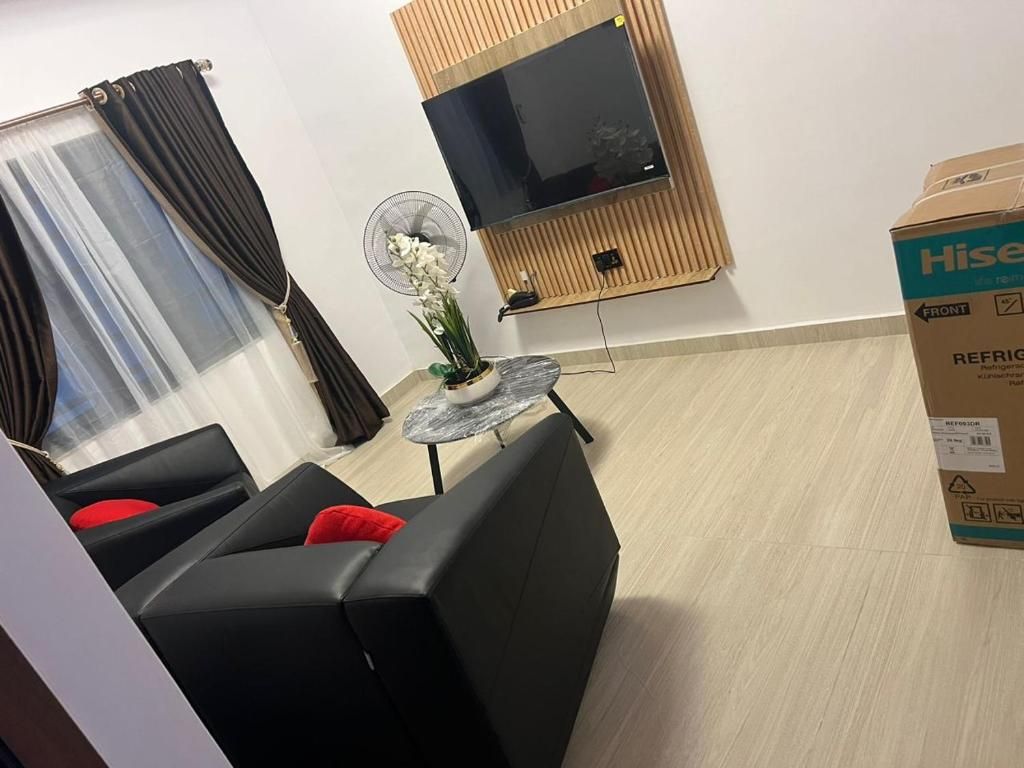 Una sala de estar con un sofá negro, una mesita redonda pequeña, un televisor de pantalla plana montado en un panel de madera y una caja con la etiqueta 'Hise' en el suelo.