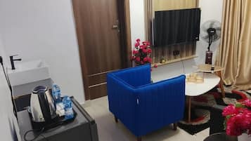 Una habitación pequeña con un sofá azul, un escritorio con un ventilador, un televisor y una puerta.