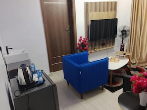 A small room with a blue sofa, a desk with a fan, a television, and a door. - De Generals Hotel (Ibadan)