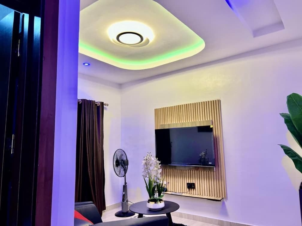 A modern living room with a flat-screen TV mounted on a wooden panel, a ceiling light fixture, and a fan.