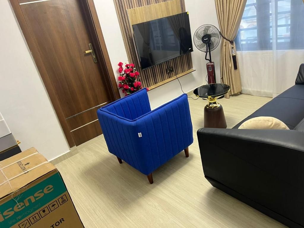 Un salón con un sillón azul, un sofá negro, un televisor, un ventilador y una ventana con cortinas.
