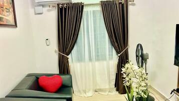 Una sala de estar con un sofá, un cojín rojo con forma de corazón, un ventilador y una orquídea en maceta.