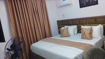 A hotel room with a bed, curtains, an air conditioner, and a fan.