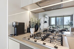 A modern kitchen with a gas stove, a coffee machine, and a view of the city through large windows. - Waterfront Escape 2BR Apt w/Pool, Gym & Parking (Docklands)