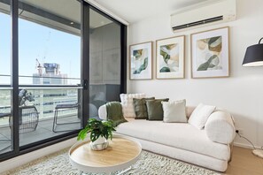 A modern living room with a large window, a sofa, a coffee table, and abstract art on the wall. - Waterfront Escape 2BR Apt w/Pool, Gym & Parking (Docklands)