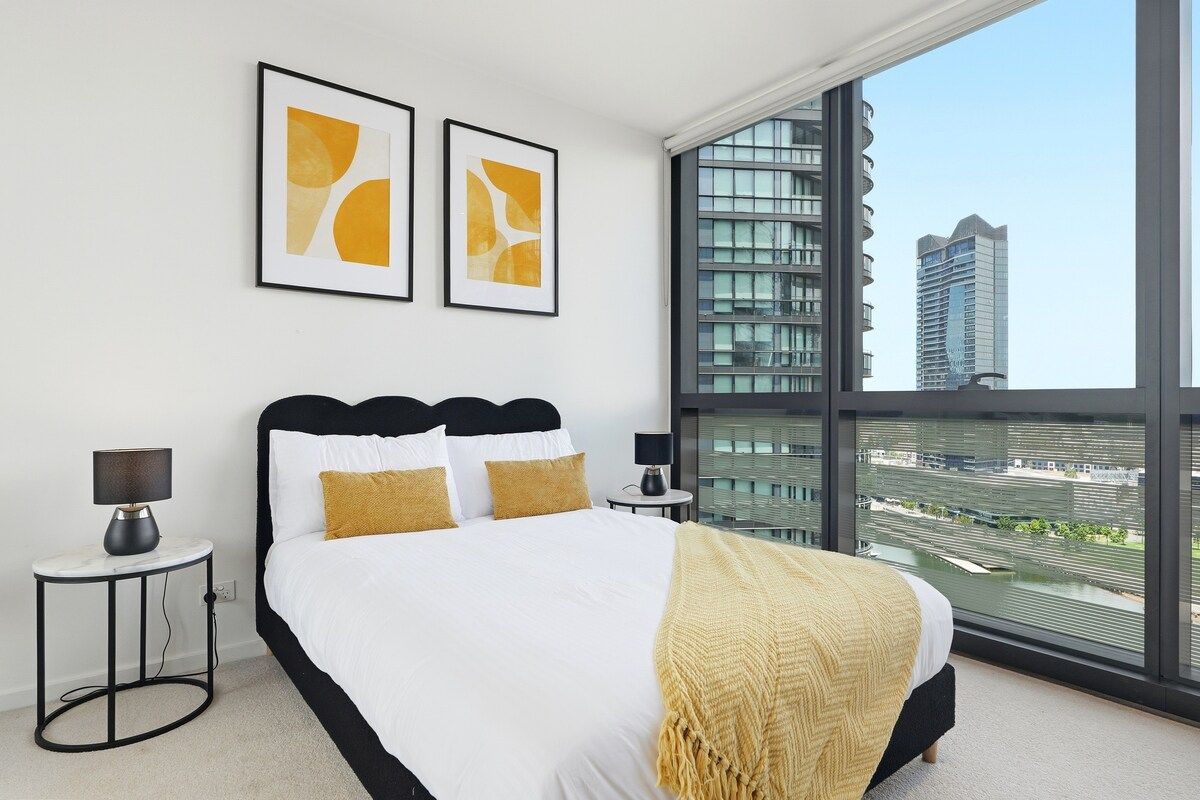 A modern bedroom with a large bed, two bedside tables, and a view of a cityscape through a large window.