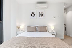 Een slaapkamer met een bed, twee nachtkastjes, een wandairconditioner en twee kunstwerken in lijsten.
