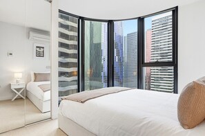 A modern bedroom with a large bed, a bedside table, a mirror, and a view of city buildings through large windows. - Highrise CBD Stay | 2B2B Apt w/Pool & Gym (Melbourne)