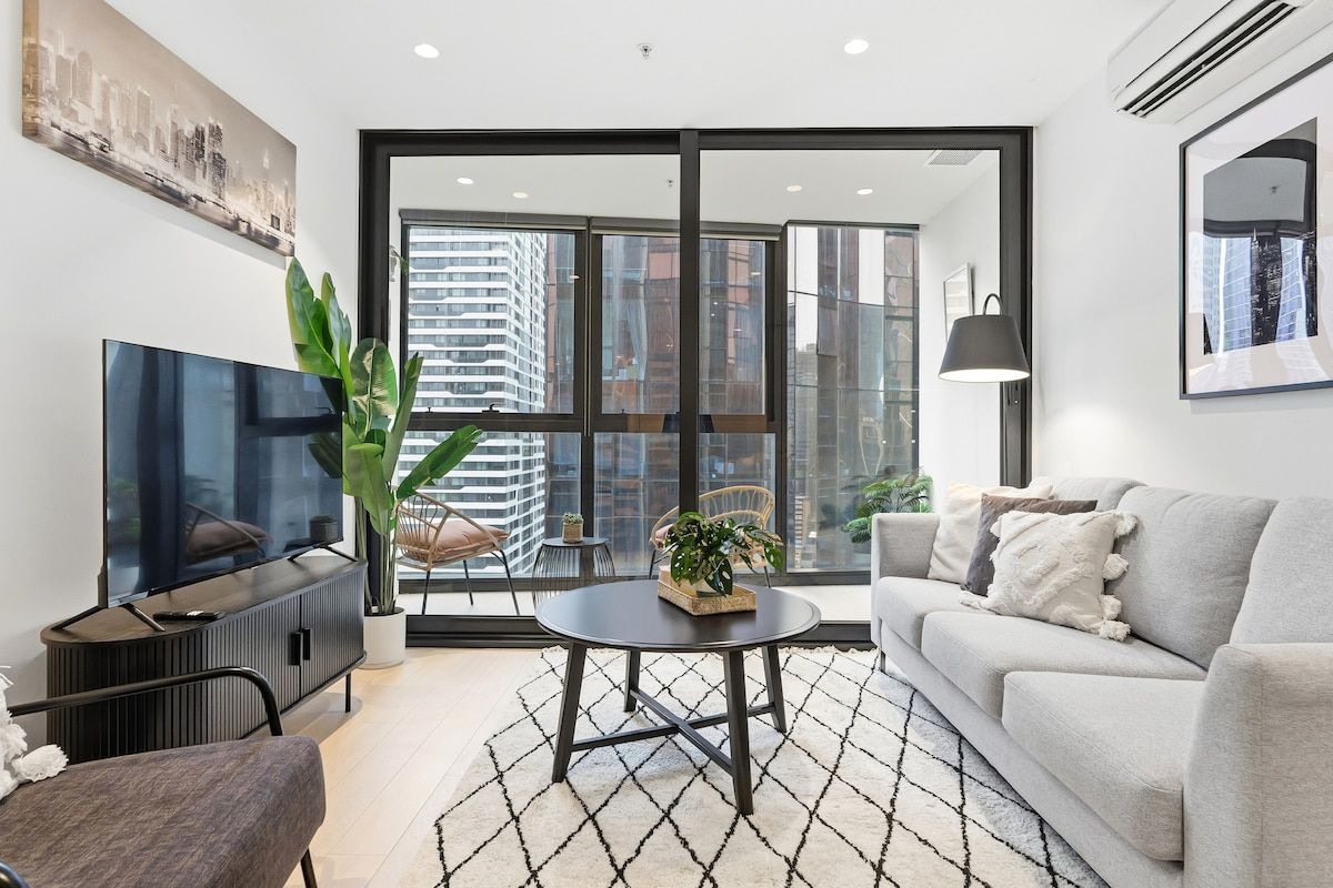 A modern living room with a flat-screen TV, a sofa, a coffee table, and a large window with a city view.