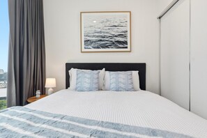 Ein Hotelzimmer mit einem Bett, einem Nachttisch, einer Lampe, einem Bilderrahmen mit einem Meerblick und Ausblick auf die Stadt.