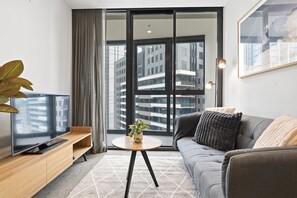 Ein modernes Wohnzimmer mit einer Couch, einem Couchtisch, einem Fernseher auf einem Holzschrank, einer Pflanze und einem großen Fenster mit Blick auf Gebäude.