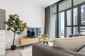 A modern living room with a large window, a flat-screen TV, a wooden sideboard, a grey sofa, and a potted plant. - Cosy 2BR apartment close to Melbourne Exhibition Centre and Crown (Southbank)