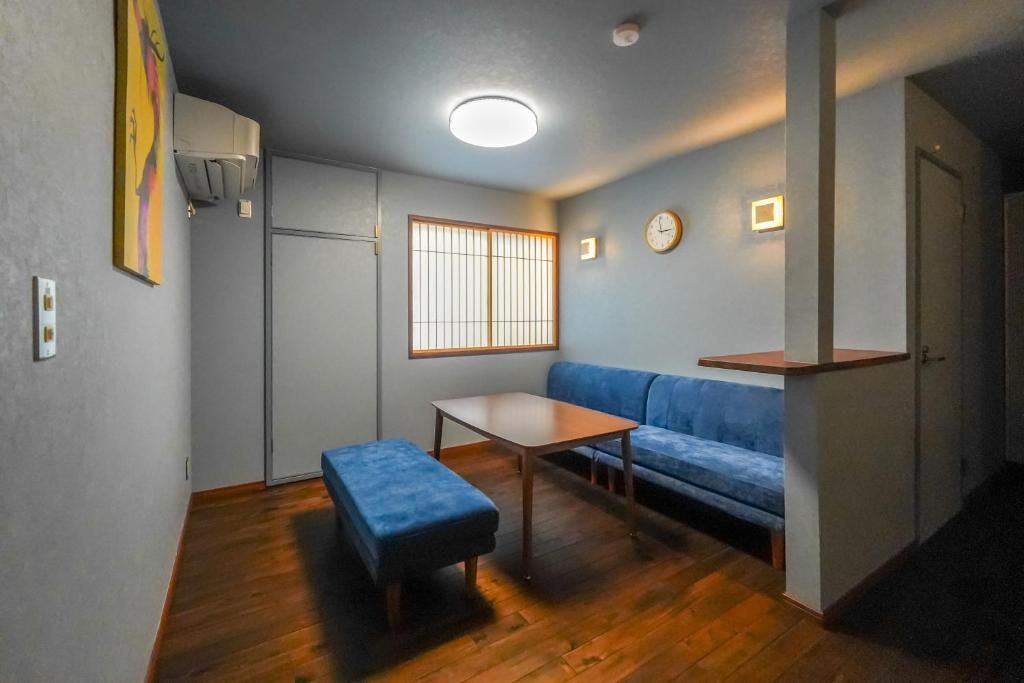 Un espacio habitable compacto con un sofá, una mesita, un banco, una ventana con persianas verticales y un reloj en la pared.