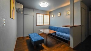 A compact living space with a sofa, a small table, a bench, a window with vertical blinds, and a clock on the wall. - Dragon Inn Nikko (nikko)