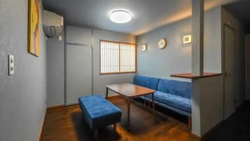 Un espacio habitable compacto con un sofá, una mesita, un banco, una ventana con persianas verticales y un reloj en la pared.