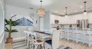 A modern kitchen with white cabinetry, stainless steel appliances, and a dining area with a glass table and white chairs. - 4BR Home with Fenced Yard & Grill (Panama City Beach)
