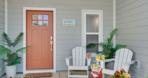 Uma casa com porta marrom, uma placa com a inscrição 'The Coastal Cove Beach House', duas cadeiras Adirondack brancas e plantas em vasos.