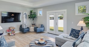 A modern living room with a flat-screen TV, two blue armchairs, a grey sofa, a coffee table with a bowl of popcorn, and a large window with a view of a green lawn. - 4BR Home with Fenced Yard & Grill (Panama City Beach)