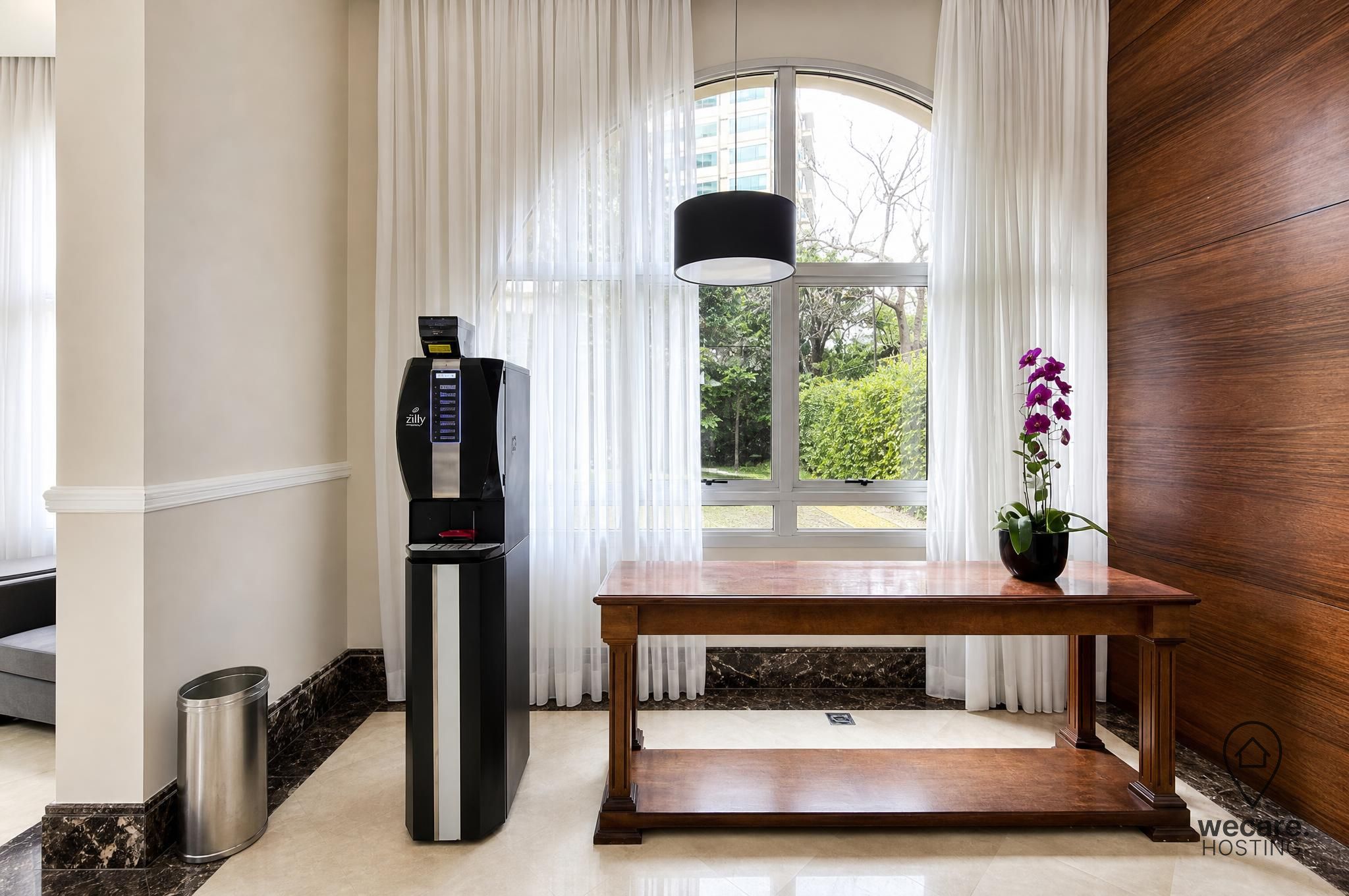 A hotel room with a minibar, a wooden table, a potted orchid, and a view of greenery through the window.