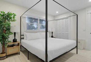 A room with a four-poster bed, a bedside table with a lamp, a potted plant, and a window with blinds. - Steps from Beach & Trolley|Heated Pool&Spa Duplex (Holmes Beach)
