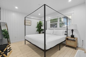 A modern bedroom with a four-poster bed, a bedside table with a lamp, a mirror, and a window with blinds. - Steps from Beach & Trolley|Heated Pool&Spa Duplex (Holmes Beach)