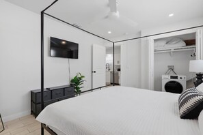 A modern bedroom with a four-poster bed, a flat-screen TV mounted on the wall, a bedside table with a plant, and a walk-in closet with a washing machine. - Main St Stunner Steps to Beach, Pool & Spa Duplex (Holmes Beach)