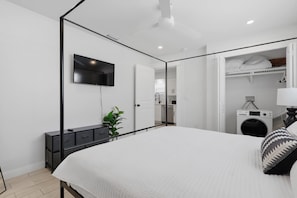 Ein modernes Schlafzimmer mit Himmelbett, einem Wand-Fernseher, einem Nachttisch mit Pflanze und einem begehbaren Kleiderschrank mit Waschmaschine.