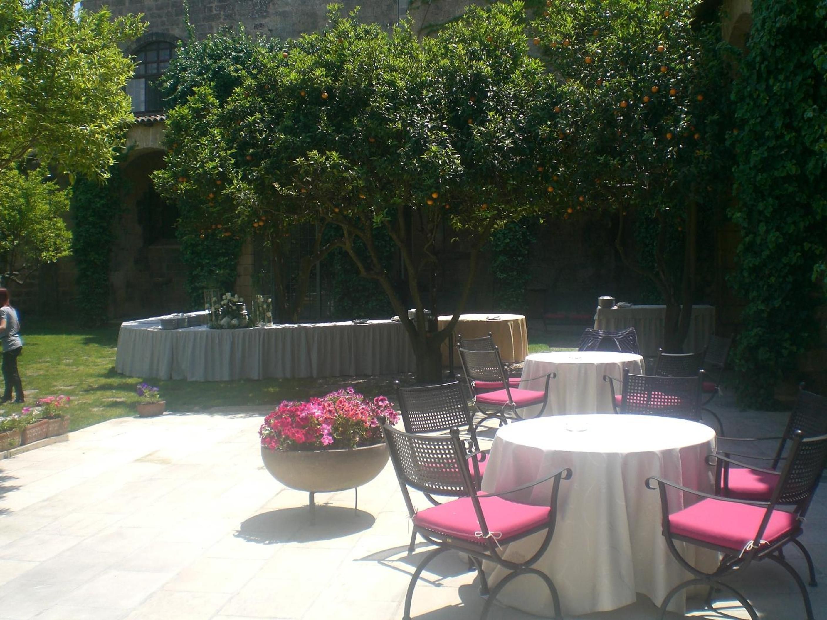 An outdoor patio with round tables and chairs, surrounded by greenery and a stone wall.