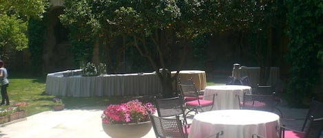 An outdoor patio with round tables and chairs, surrounded by greenery and a stone wall.