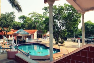 A swimming pool with a statue, surrounded by trees and a covered area with tables and chairs.