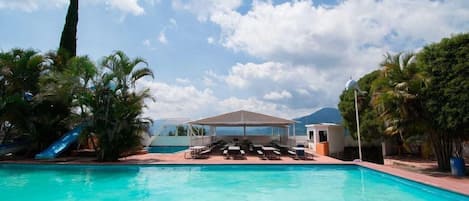 A swimming pool with lounge chairs and a shaded area, surrounded by palm trees and a clear sky.