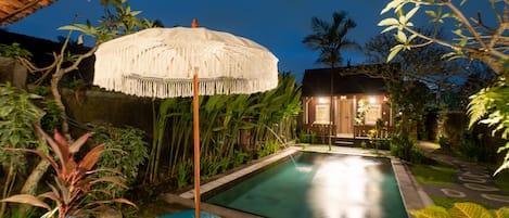 A swimming pool with a white umbrella, surrounded by tropical plants and a wooden structure in the background.