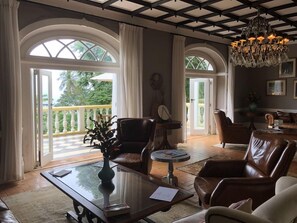 A spacious living room with a large window, a chandelier, leather furniture, and a view of greenery outside. - Belo Monte Hotel and Museum (Príncipe Island)