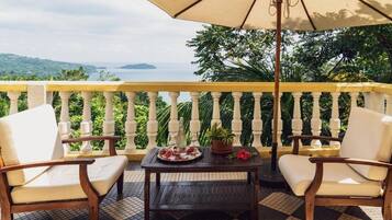 Ein Balkon mit Schachbrettboden, zwei weiße Sitzgelegenheiten mit Kissen, ein Holztisch mit Blumenarrangement und ein großer Sonnenschirm.