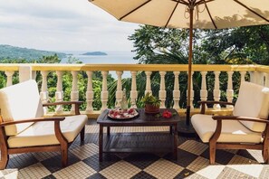 A balcony with a checkered floor, two white cushioned chairs, a wooden table with a floral arrangement, and a large umbrella. - Belo Monte Hotel and Museum (Príncipe Island)