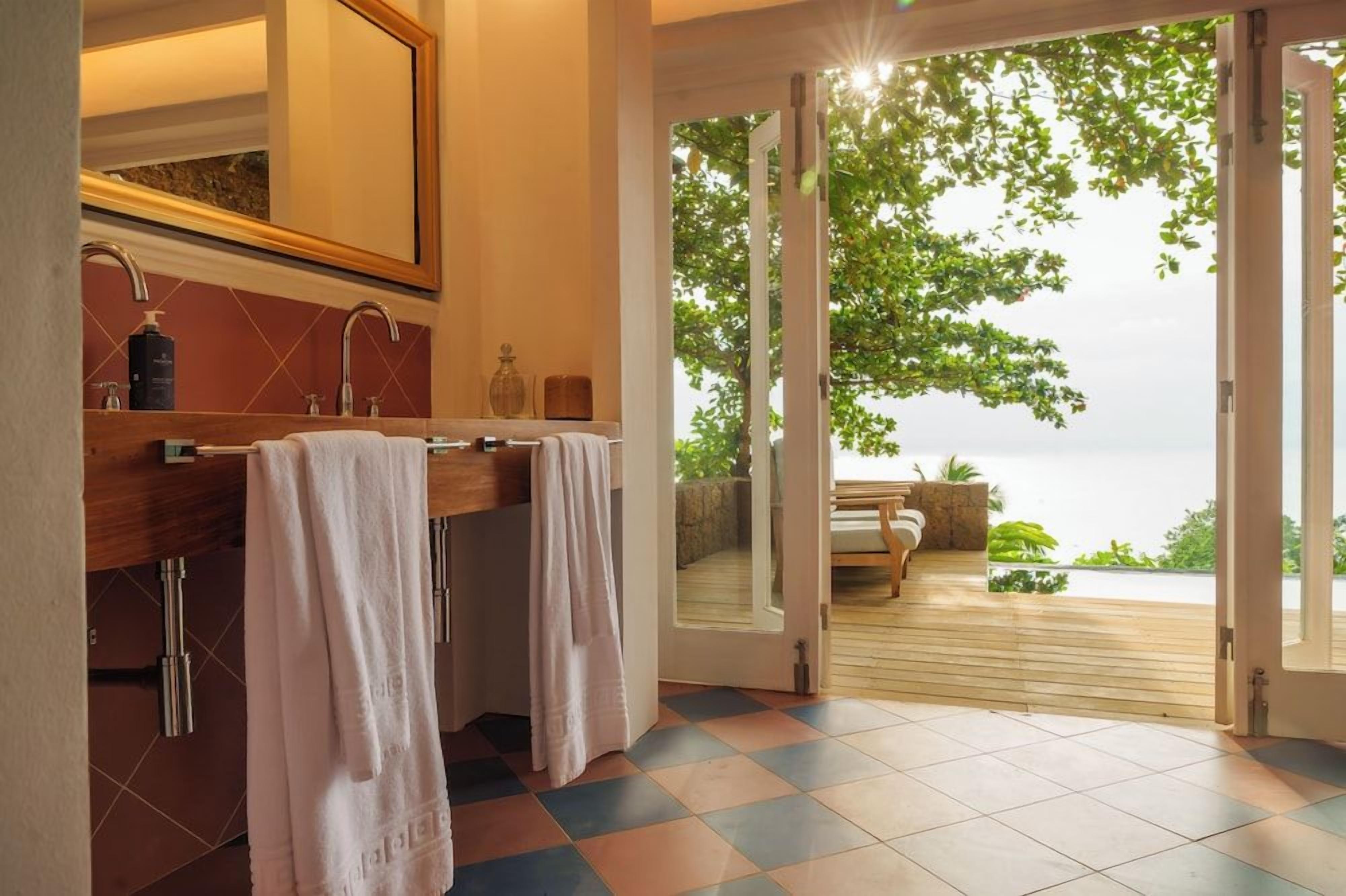 A bathroom with a large mirror, two sinks, and a view of an outdoor deck with a bed and greenery.
