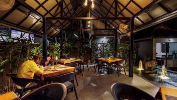 An indoor dining area with tables and chairs, surrounded by plants and a water feature.