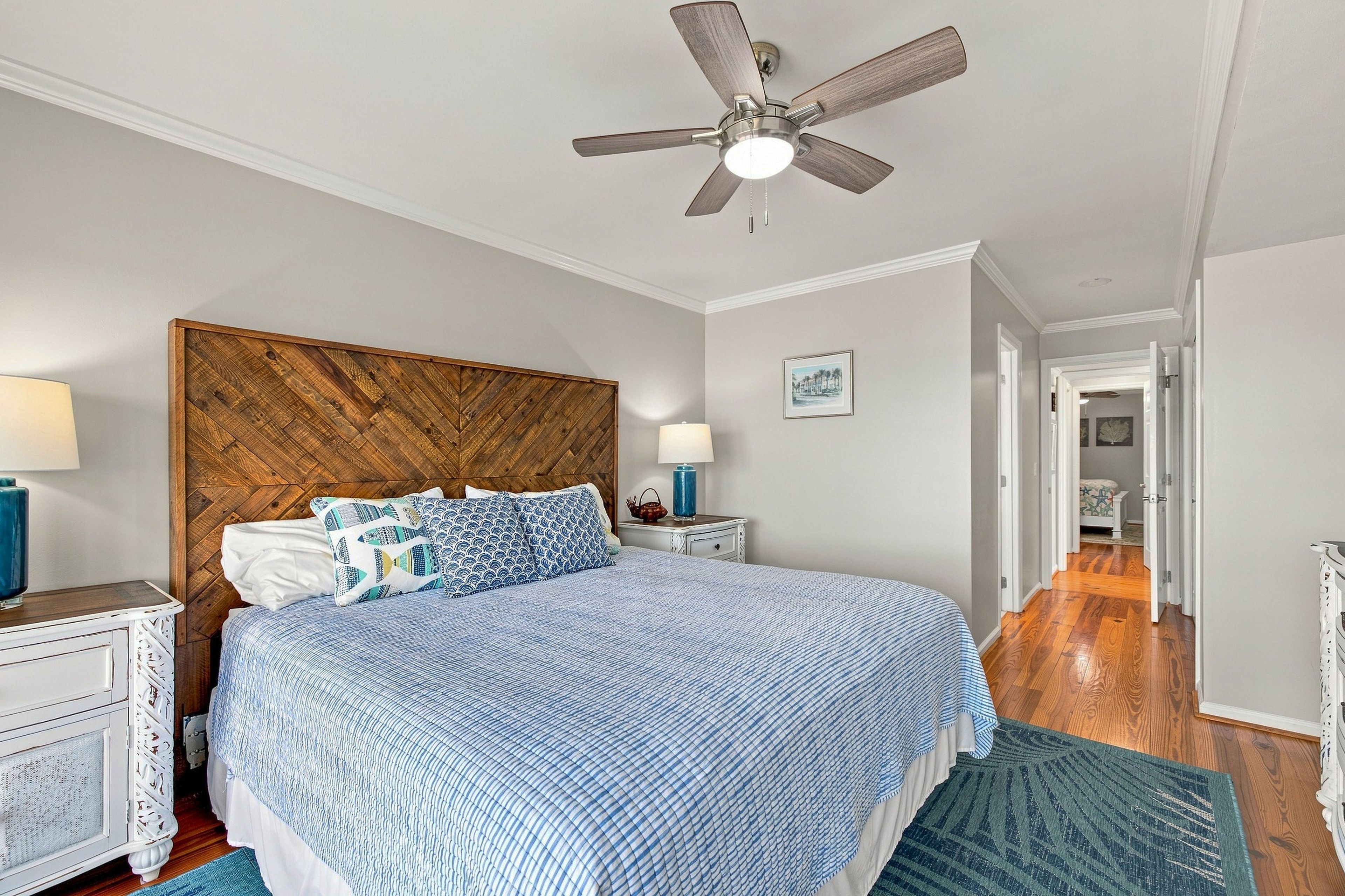 Quarto com cama, cabeceira de madeira, ventoinha de teto, mesa de cabeceira, luminária e porta que dá para outro cômodo.